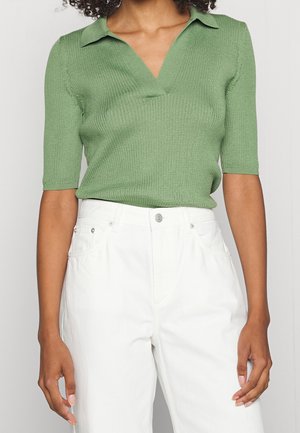 Person iført en ribbet, lysegrøn striktop med krave og korte ærmer, stoppet ned i højttaljede hvide bukser med forlommer og knaplukning.