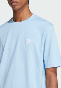 Tricou din bumbac de culoare albastru deschis, cu mâneci scurte și guler rotund, având un mic logo Adidas alb imprimat pe piept.