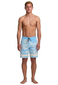 Muž stojaci naboso, oblečený v svetlomodrých plážových šortkách so svetlými vodorovnými pruhmi a čiernou šnúrkou, s neutrálnym výrazom.