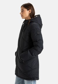 Svart isolert parka med hette, med kviltet design, glidelåslommer og et logo-patch på ermene. Myk innvendig fôr.