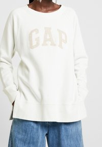 Sudadera crema con el logo "GAP" en beige. Cuenta con mangas raglán, un cuello ancho y un corte relajado. Tejido suave y texturizado.