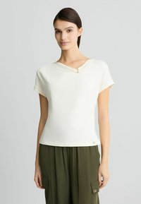 Krem-bluza kratkih rukava s drapiranim dekolteom, s zlatnim detaljem blizu ovratnika i logom na rubu.