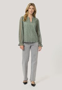 Blusa verde chiaro con motivo geometrico, maniche lunghe in tessuto trasparente, scollatura a V e polsini arricciati, abbinata a jeans grigi chiari e tacchi neri.