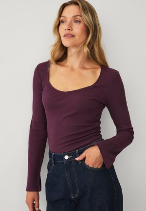 Kvinde med langt blond hår iført en tætsiddende langærmet lilla top med bådudskæring og mørkeblå højtaljede jeans, stående mod en hvid væg.