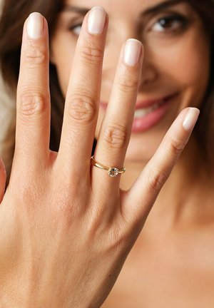 Gold ring with a round, clear gemstone resting on a hand with light skin and neutral nail polish, displayed prominently against a blurred background.