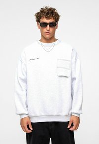 Sweatshirt gris clair avec une poche avant, col rond, manches larges et un discret logo brodé noir. Porté avec des lunettes de soleil.