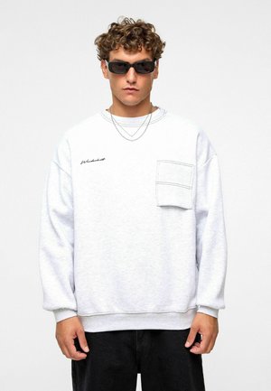 Lichtgrijze sweatshirt met een voorkraag, ronde hals, brede mouwen en subtiele zwart geborduurde branding. Gecombineerd met een zonnebril.
