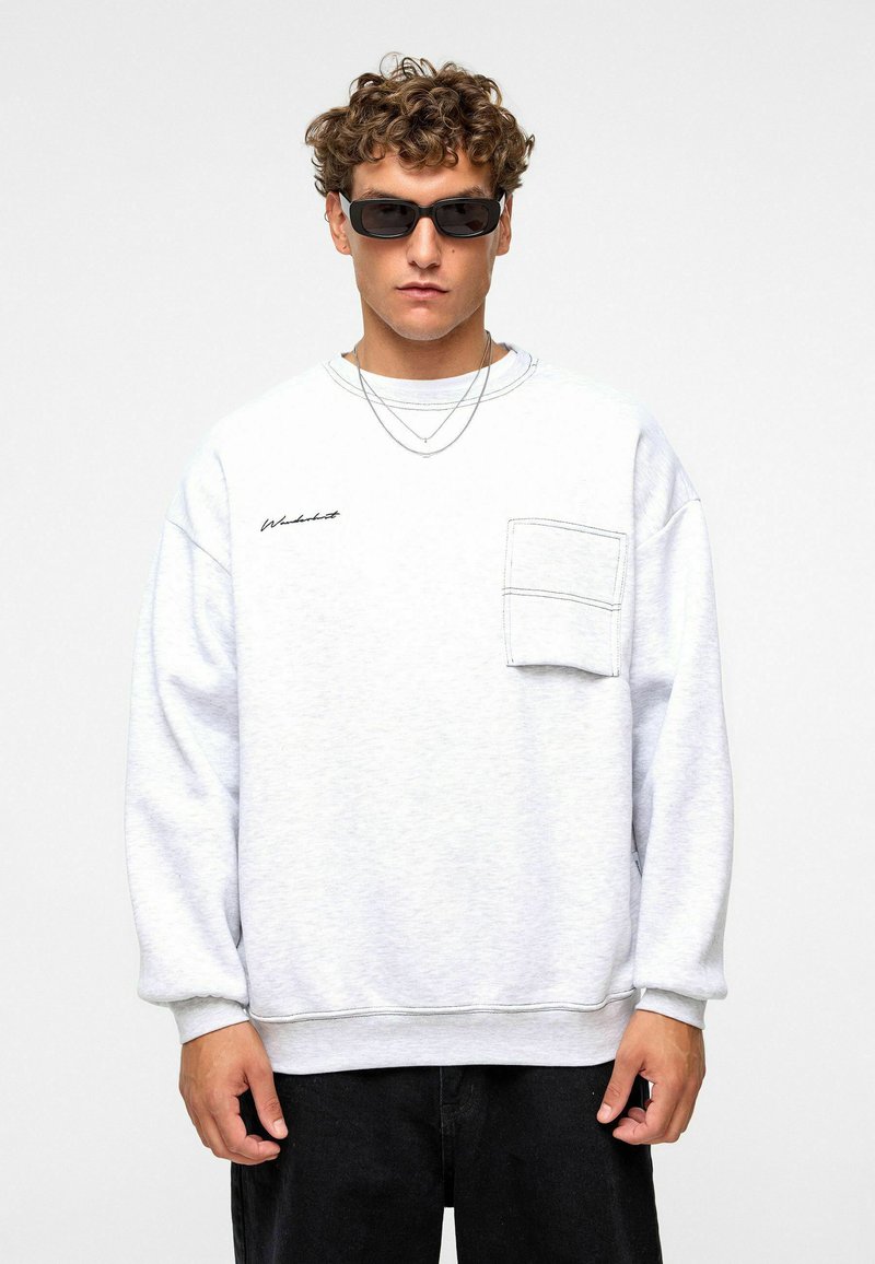 Sweatshirt gris clair avec une poche avant, col rond, manches larges et un discret logo brodé noir. Porté avec des lunettes de soleil.
