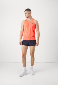Neon-orange Sporttanktop mit einem strukturierten geometrischen Muster, kombiniert mit dunkelblauen Shorts. Das Model trägt weiße Sneaker und Knöchelsocken.