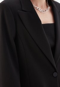 Svart skräddarsydd kavaj med slagkrage, knappdetalj och en mjuk textur, kombinerad med ett silverfärgat kedjehalsband.