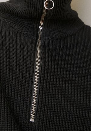 Zbliżenie czarnego swetra z prążkowanego dziania z srebrnym metalowym suwakiem i okrągłym uchwytem przy szyi.