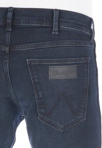 Vaqueros de mezclilla azul oscuro con corte recto, que presentan un parche de cuero con el logo de Wrangler en el bolsillo trasero y detalles de costura en contraste.