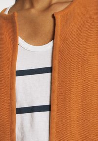 Gros plan d'une personne portant une chemise blanche avec deux rayures horizontales noires et un cardigan orange texturé ouvert.