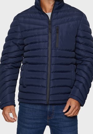 Homme portant une doudoune matelassée bleu marine avec des poches zippées à l'avant et sur la poitrine, associée à un jean bleu.