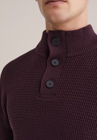 Bordeaux gebreide pullover met een hoge kraag en drie zwarte knopen. Geweven stof met een wafelpatroon. Close-up van de halslijn.