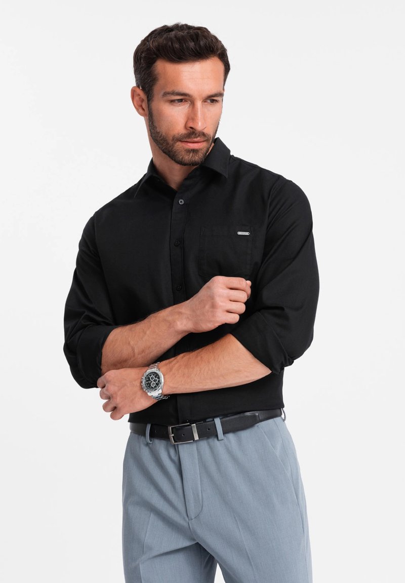 Zwarte overhem leefje met lange mouwen, slim fit, voorzak, gecombineerd met lichtgrijze broeken en een zilveren horloge. Effen structuur.