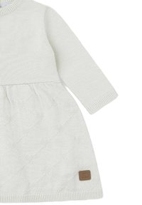 Vestido blanco de punto para bebé con mangas largas, patrón de diamantes texturizado y pequeño parche de logo marrón cerca del dobladillo.