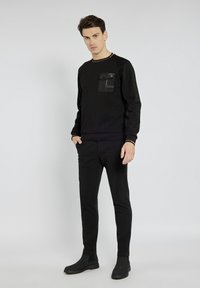 Czarna sweter z prążkowanej tkaniny z kieszenią na klatce piersiowej, kontrastującym żółtym dekoltem oraz dopasowanymi czarnymi spodniami slim-fit i butami. Gładka tekstura, minimalistyczny design.