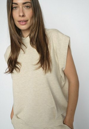 Young woman with long brown hair wearing a beige sleeveless top, standing with hands in pockets against a plain light background.