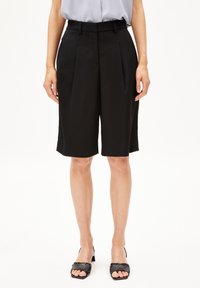 Schwarze maßgeschneiderte Shorts mit Falten, aus leichtem Stoff gefertigt. Mittellang, mit einem Bund und glatter Textur. Tragen mit schwarzen offenen High Heels.