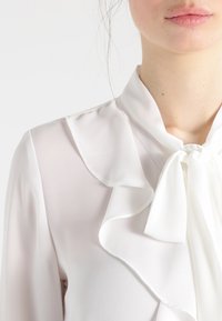 Blusa bianca realizzata in tessuto leggero, con colletto arricciato e dettaglio a fiocco al collo, con una texture liscia e leggermente trasparente.