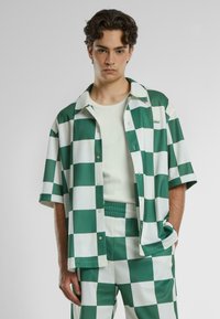 Camicia a quadri verde e bianca con maniche corte, chiusura a bottoni e colletto rilassato, abbinata a pantaloni a quadri coordinati.