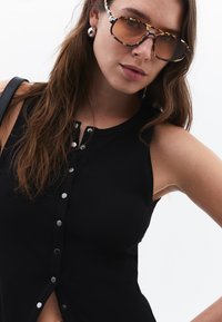 Top sans manches en maille noire avec des boutons-pression en métal sur le devant. Le modèle porte des lunettes en écaille de tortue marron et des boucles d'oreilles en argent.