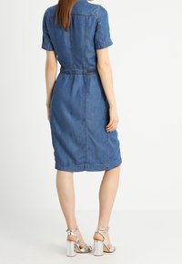 Abito in denim con maniche corte, vita strizzata, taglio al ginocchio e una consistenza liscia. Presenta una cucitura posteriore e dettagli cuciti. Tacchi argento visibili.