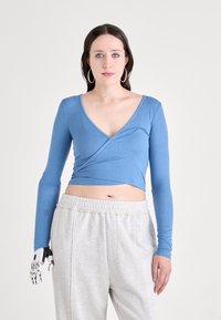 Svijetloplava dugosleeve wrap majica s V-izrezom, napravljena od rebraste tkanine. Uparen sivi trenirka s teksturiranom gumicom.