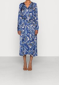 Frau, die ein langes blaues Blumen-Wickelkleid mit langen Ärmeln und einem V-Ausschnitt trägt, kombiniert mit braunen, offenen Sandalen mit Riemen.