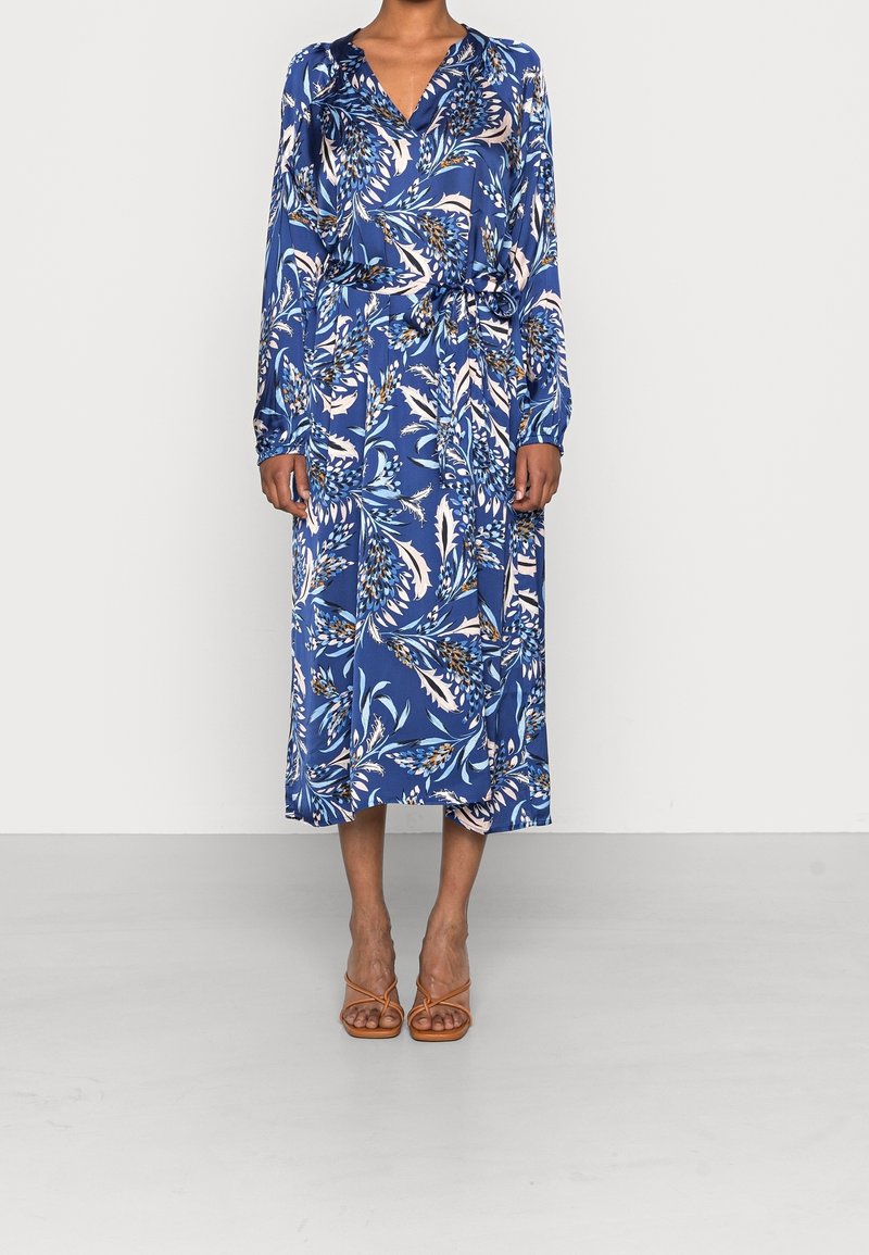 Frau, die ein langes blaues Blumen-Wickelkleid mit langen Ärmeln und einem V-Ausschnitt trägt, kombiniert mit braunen, offenen Sandalen mit Riemen.