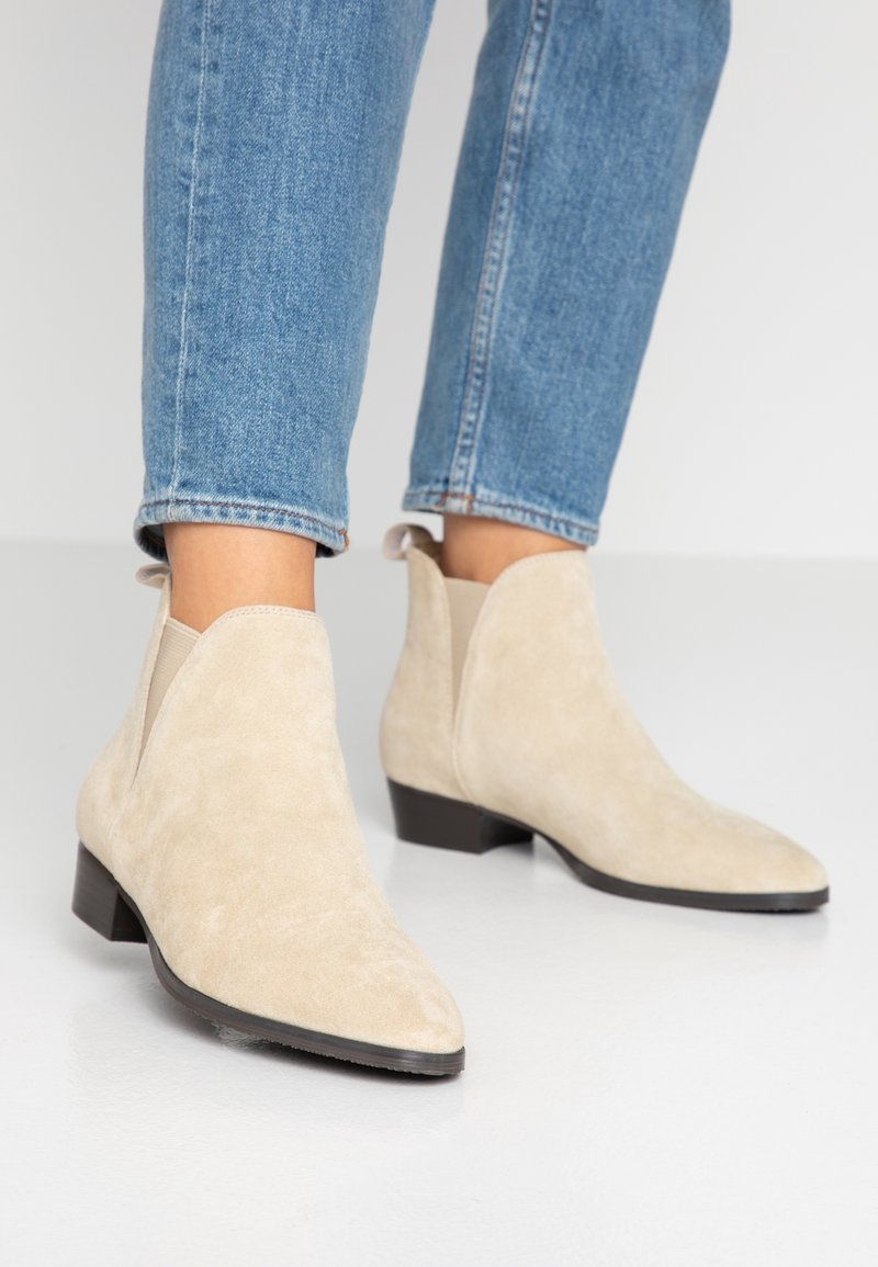Beige mokkasiner med spiss tå og lav blokkheel, med elastiske sidepaneler og en glidelås bak.