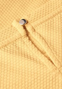 Žlutá texturovaná tkanina s vzorem včelí plástve, se zapínáním na metalické patentky a viditelným prošitím.