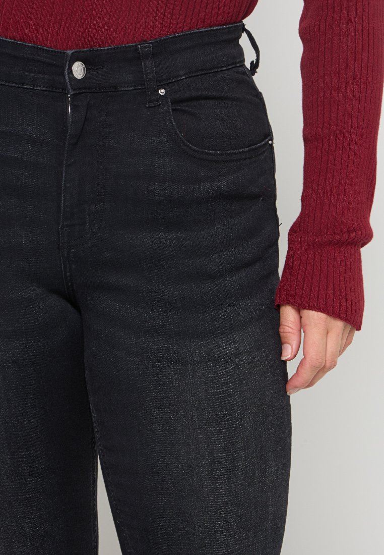 Schwarze High-Waist-Jeans mit schmaler Passform, ausgestattet mit einer Vorderseitentasche, silbernem Zubehör und einem leichten Glanz. Kombiniert mit einem strukturierten burgunderroten Pullover.