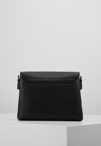 Černá kožená obdélníková kabelka přes rameno s klopou a zlatými kovovými detaily na stranách, umístěná na bílé ploše na světlém šedém pozadí.