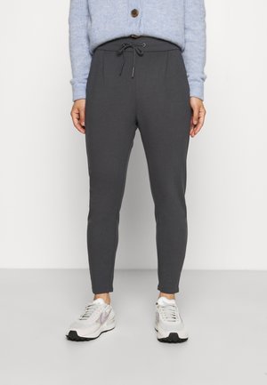 Person iført mørkegrå joggebukser med snøre, lyseblå cardigan og hvide sneakers, stående på en simpel lysegrå gulv.