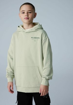 Niño con sudadera con capucha de color verde claro de AllSaints con bolsillo frontal y pantalones negros, sobre un fondo gris liso.
