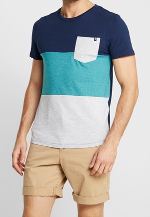 Mann trägt ein T-Shirt in Farbabblockung mit Marineblau, Türkis und Hellgrau und einer Brusttasche, kombiniert mit hochgekrempelten beigen Shorts.