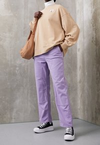 Felpa oversize beige con logo, pantaloni viola chiaro con tasche laterali e sneakers nere con suola bianca. Borsa a tracolla marrone.