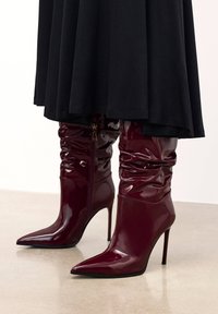 Bottes hautes brillantes bordeaux décontractées jusqu'au genou, avec des bouts pointus et des talons aiguilles, portées sous une jupe noire.