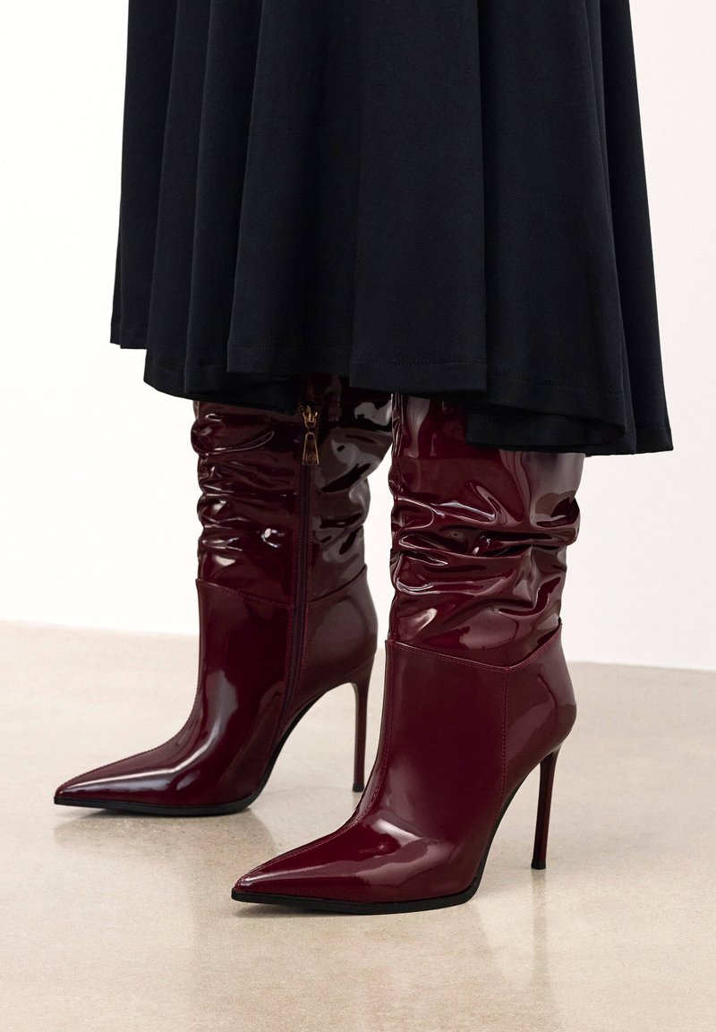 Bottes hautes brillantes bordeaux décontractées jusqu'au genou, avec des bouts pointus et des talons aiguilles, portées sous une jupe noire.