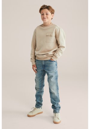 Beżowy sweatshirt z kieszenią, zestawiony z jasnoniebieskimi dżinsami. Białe buty z beżową podeszwą. Neutralne tło.