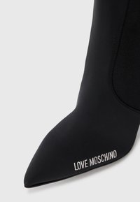 Sorte ankelstøvler i stoff med spiss tå, med teksturert design og hvit "LOVE MOSCHINO" branding på siden.