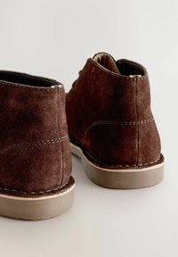 Scarpe in suede marrone con una finitura testurizzata, caratterizzate da una suola chiara in contrasto, cuciture visibili e un design a caviglia bassa.