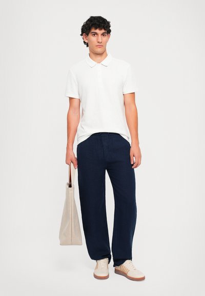 Junger Mann steht da und trägt ein weißes Poloshirt, navyblaue Hosen, beigen Sneakers und hält in seiner linken Hand eine beigen Tragetasche.