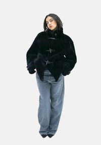 Schwarzer Kunstfellmantel mit hohem Kragen, großen Ärmeln und Knebelverschlüssen. Kombiniert mit blauen Jeanshose mit gesprenkeltem Muster.