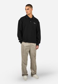 Μαύρο πόλο μπλουζάκι με μακριά μανίκια από υφασμάτινη υφή και γιακά, συνδυασμένο με ανοιχτά μπεζ σουέτ παντελόνια και λευκά αθλητικά παπούτσια.