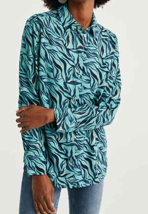 Kobieta ubrana w turkusową koszulę na guziki z niebieskim i beżowym abstrakcyjnym falistym wzorem, w parze z niebieskimi dżinsami, stojąca z założonymi ramionami.