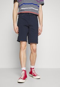 Námořnické modré šortky z bavlněného směsového materiálu, slim fit s poutky na opasek, předními kapsami a zakončením nad kolenem. Slučují se s červenými vysokými teniskami.