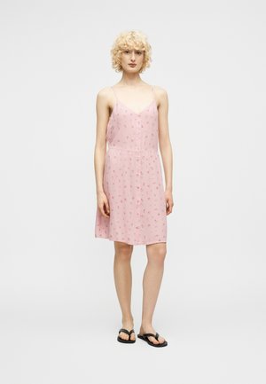 Persoon met krullend blond haar, gekleed in een mouwloze roze jurk met kleine bloemenprint en zwarte sandalen, staand tegen een effen achtergrond.